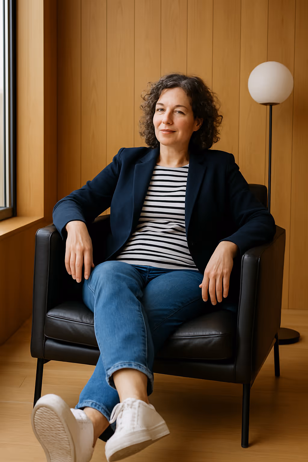 Person sitzt im Sessel, trägt dunkelblauen Blazer und Ringelshirt, lächelt leicht.

