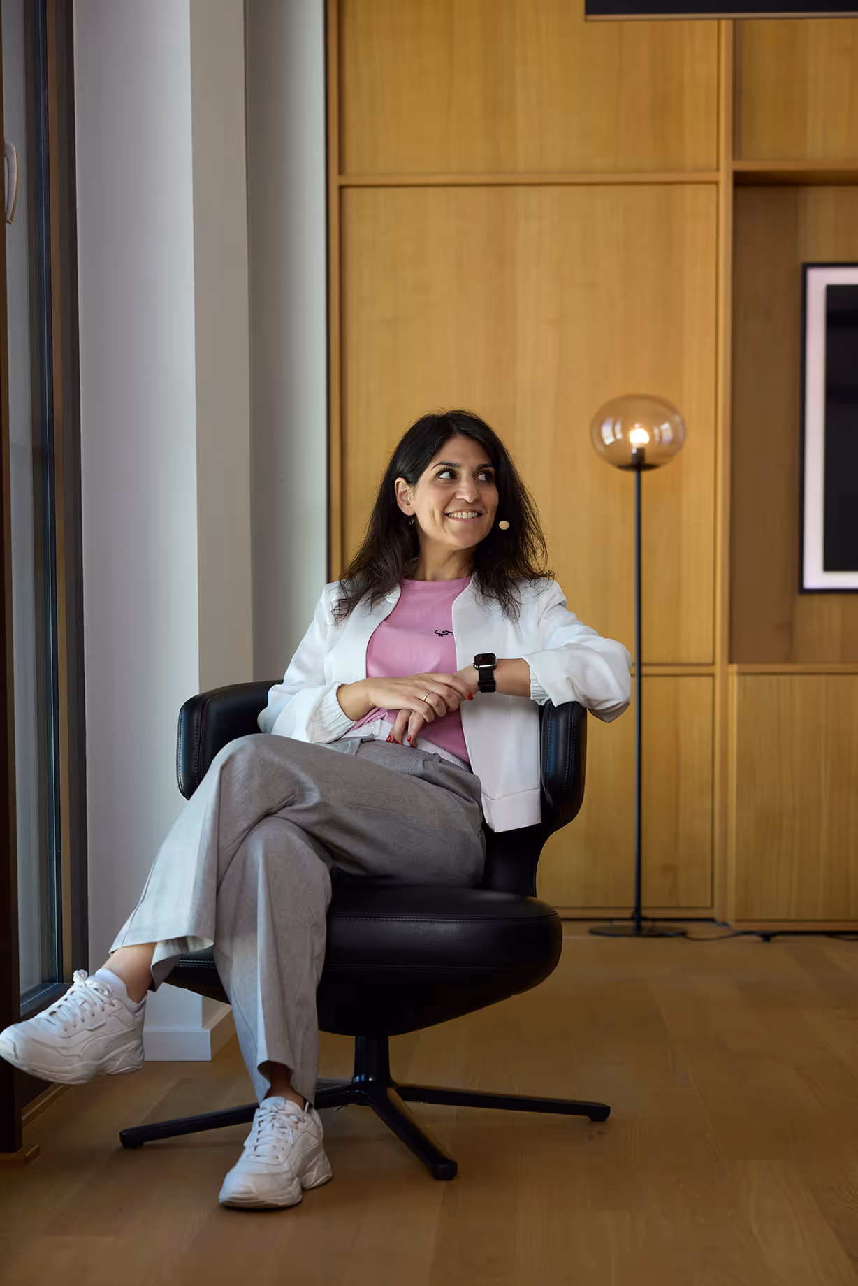 Person sitzt im Sessel, trägt weißen Blazer und rosa Shirt und lächelt zur Seite.
