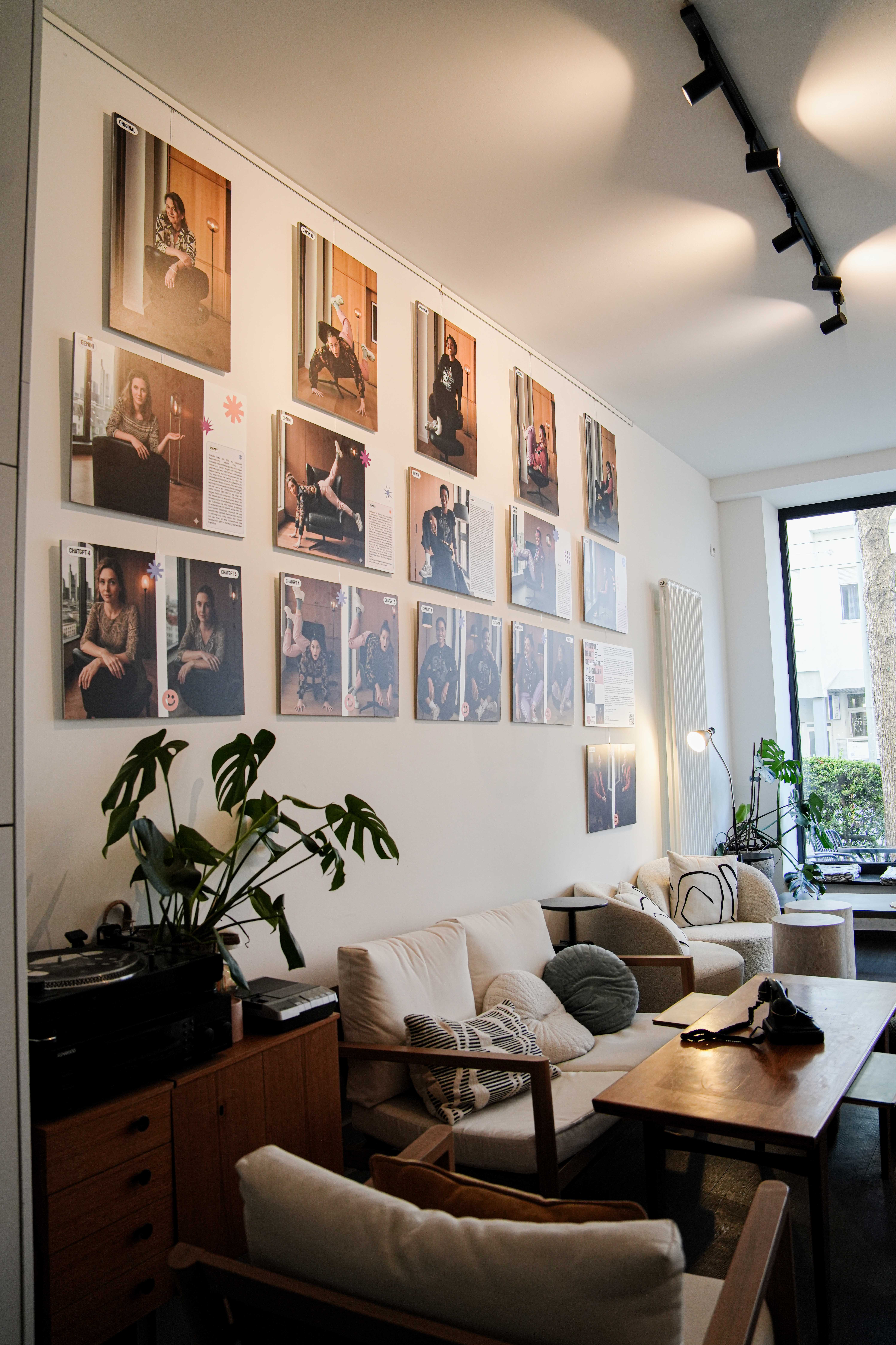 Gemütliches Wohnzimmer mit Sofa, Sesseln, Wandfotos und großen Fenstern mit Blick auf grüne Hecke.