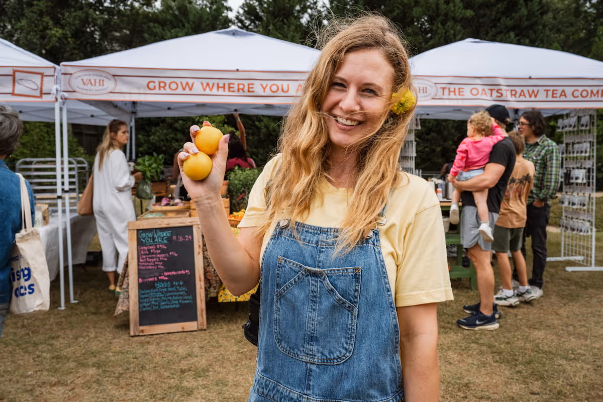 A woman in overalls with a yellow flower behind her ear, holds fresh produce in front of a row of vendors at the VaHi Farmers Market.