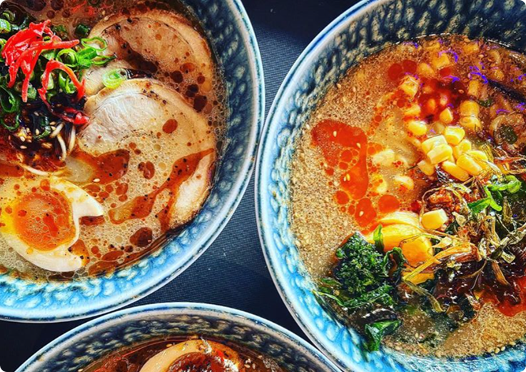 Three bowls of ramen with broth, sliced pork, half-boiled eggs, corn, greens, and chili oil.