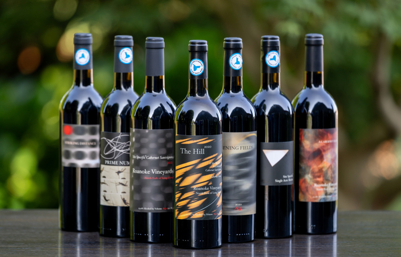 Seven wine bottles from Roanoke Vineyards arranged in a row on a wooden surface with a blurred green background.