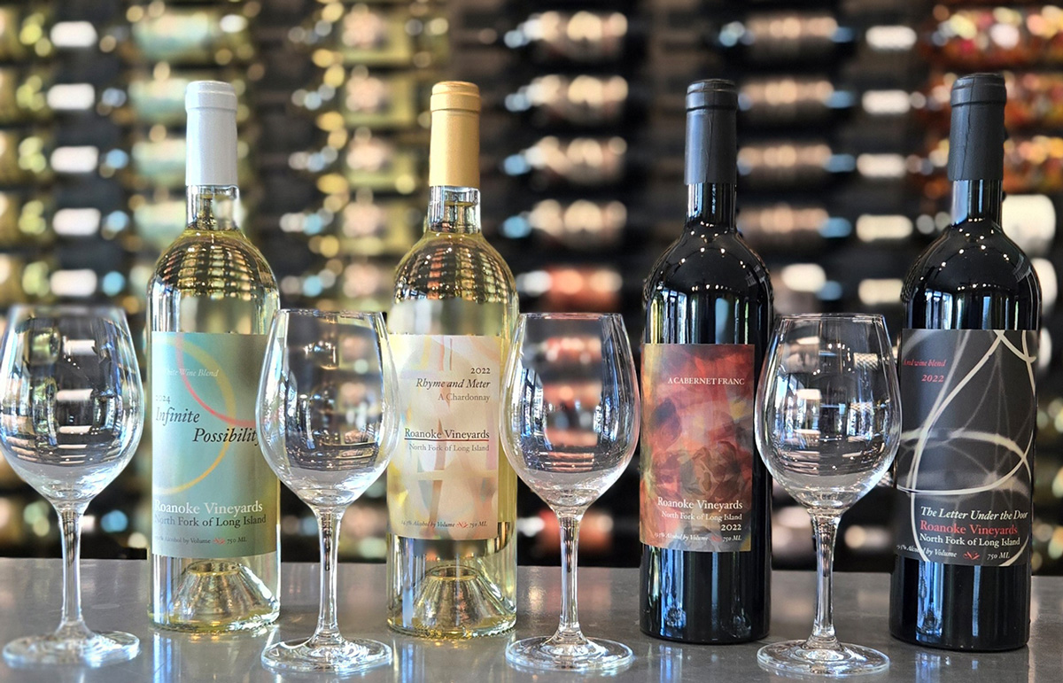 Four bottles of Roanoke Vineyards wine—two white and two red—each paired with a wine glass, arranged on a marble counter with a blurred wine rack background.
