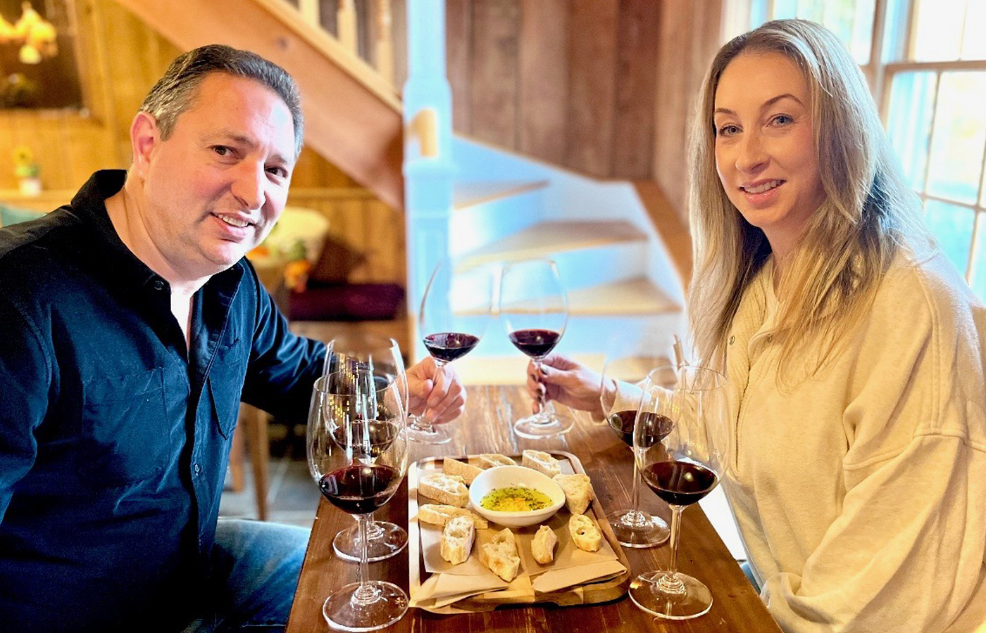 Man and woman sitting at a wooden table with glasses of red wine and a plate of bread with olive oil dip, smiling indoors.