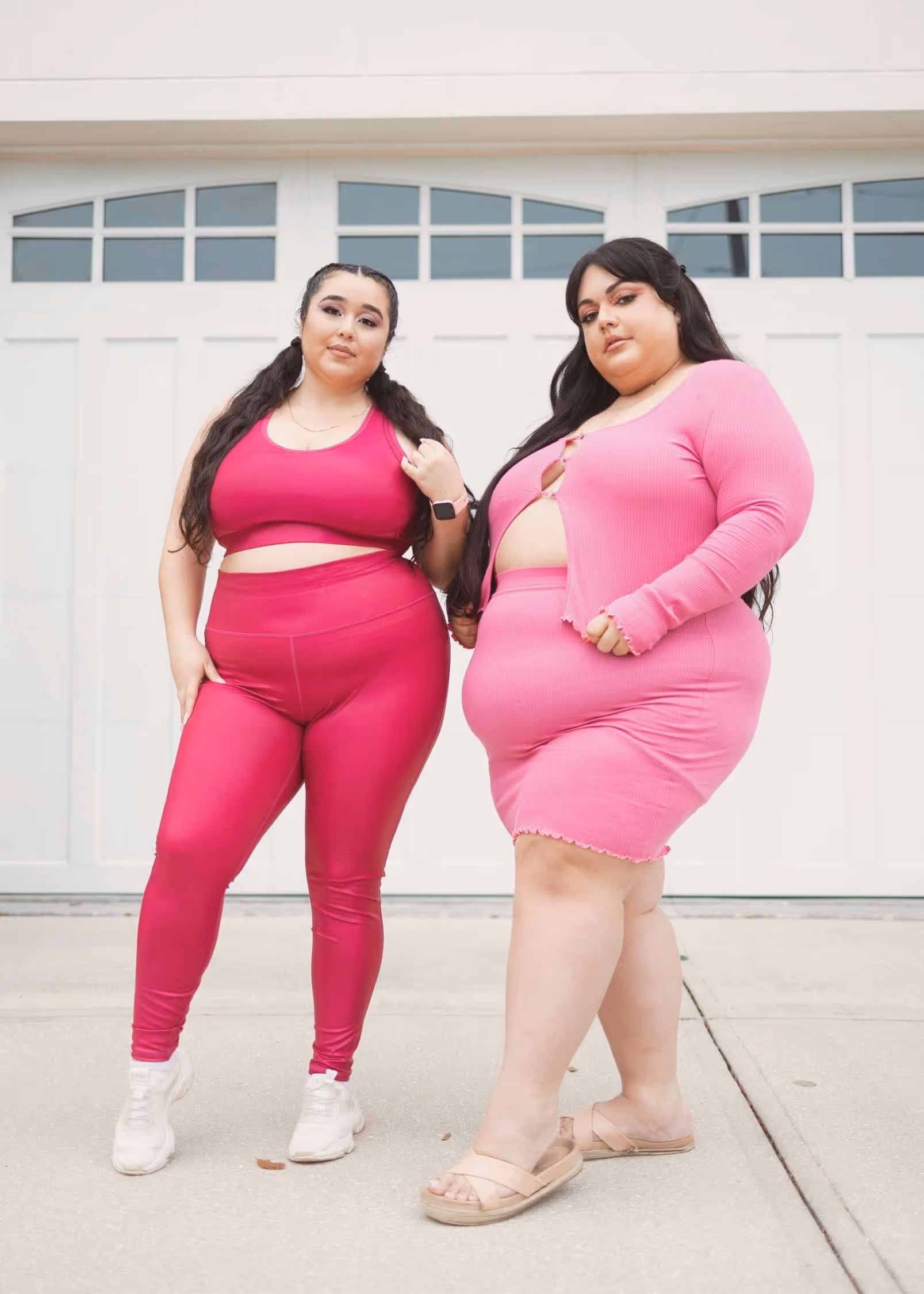 Two fat babes with long, straight, dark hair stand in front of a white garage door wearing bright pink active wear sets.
