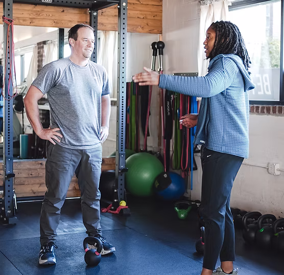 Two people in workout attire conversing in a gym with kettlebells and resistance bands visible.