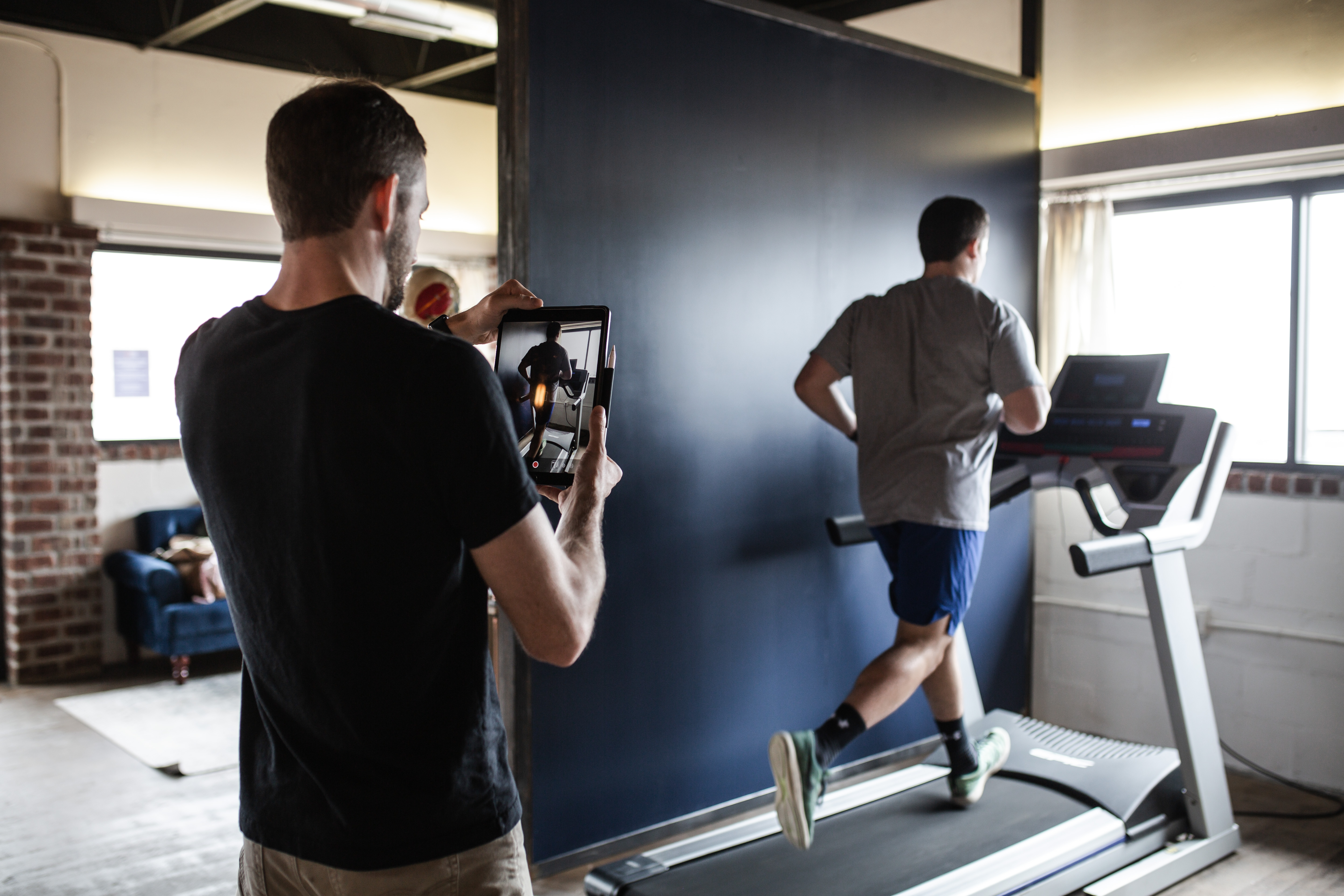 A man records another man running on a treadmill indoors with a tablet.