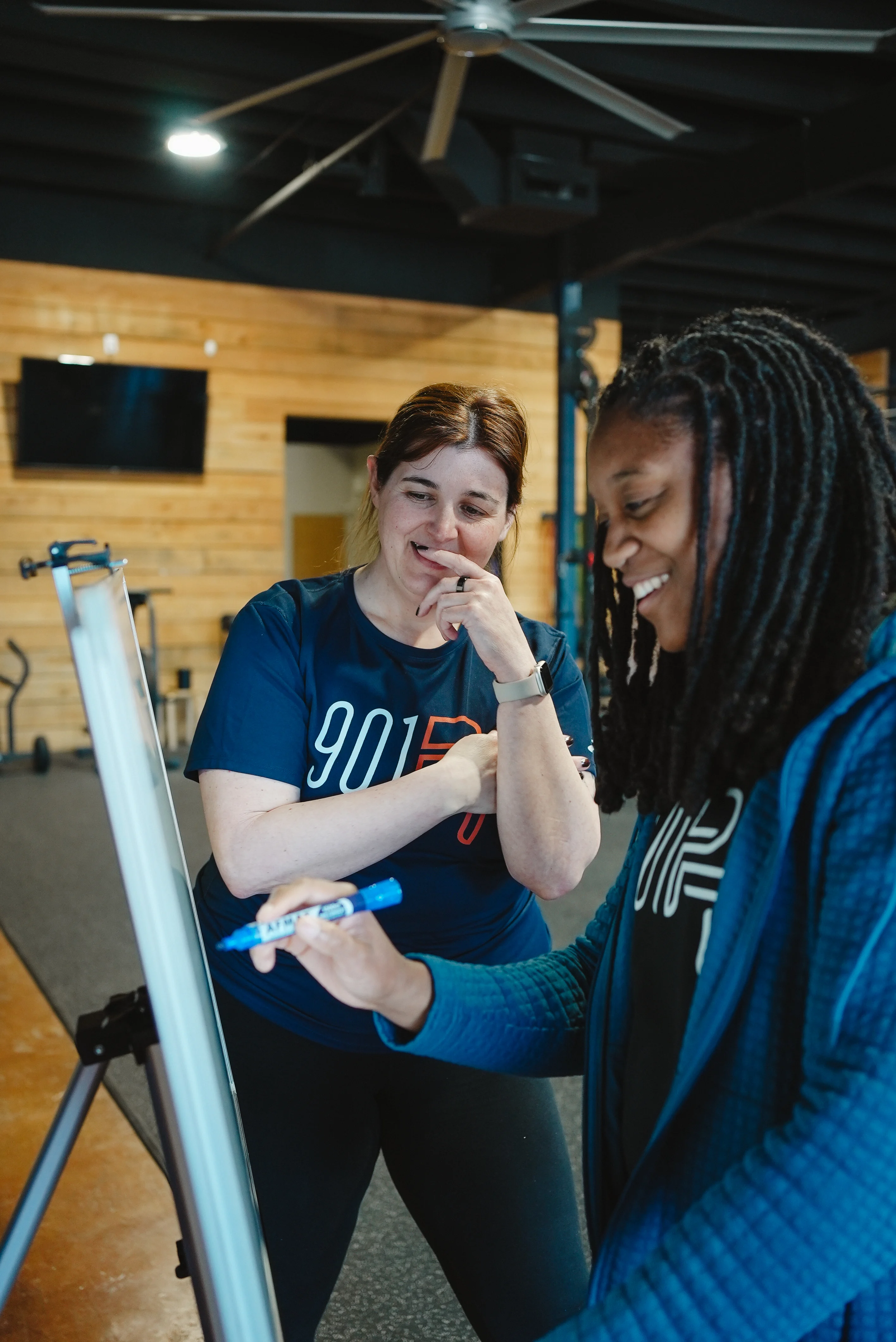 Two women collaborating and writing on a whiteboard in a modern gym setting.