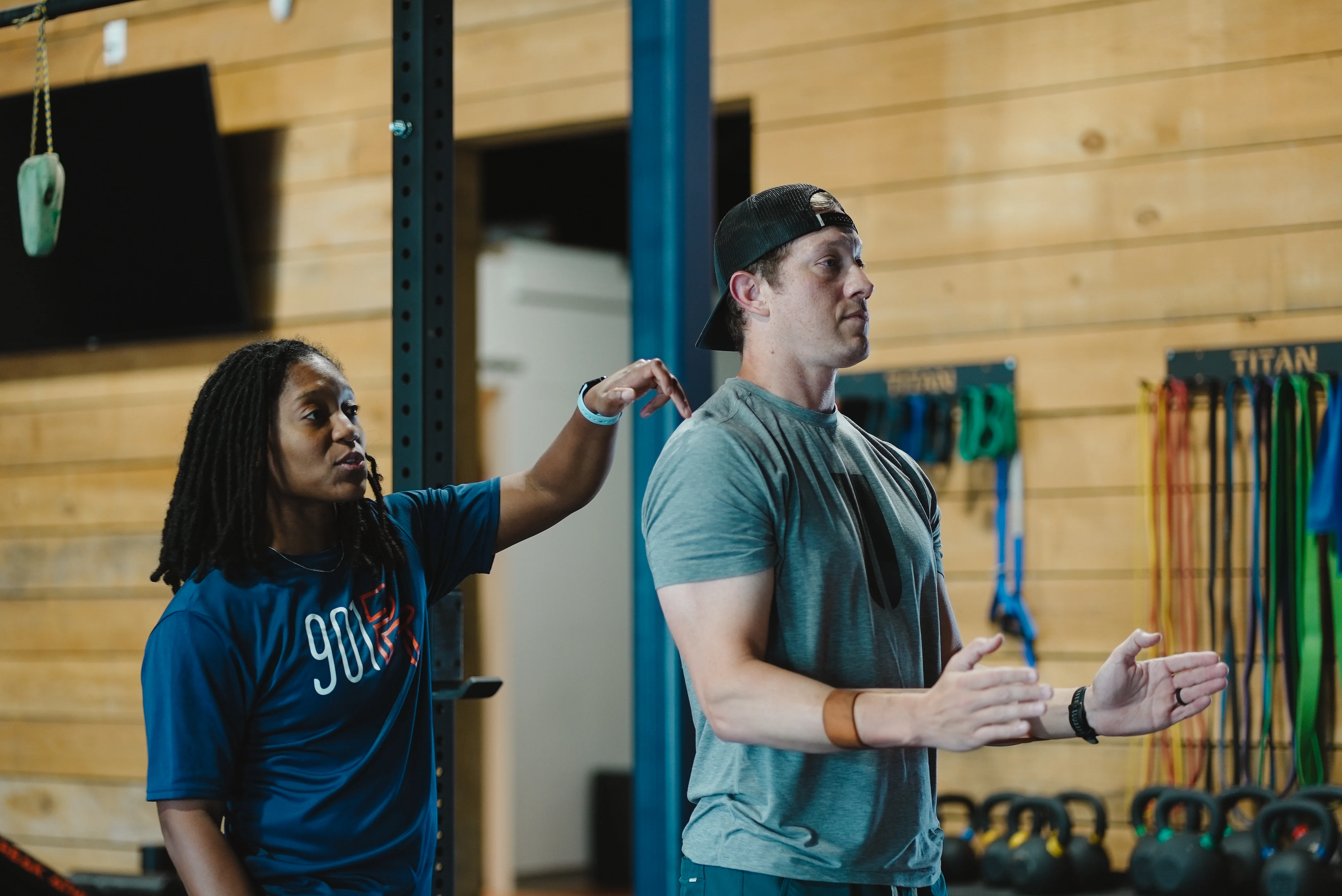 Therapist instructing a man performing an exercise in a gym with kettlebells and resistance bands in the background.