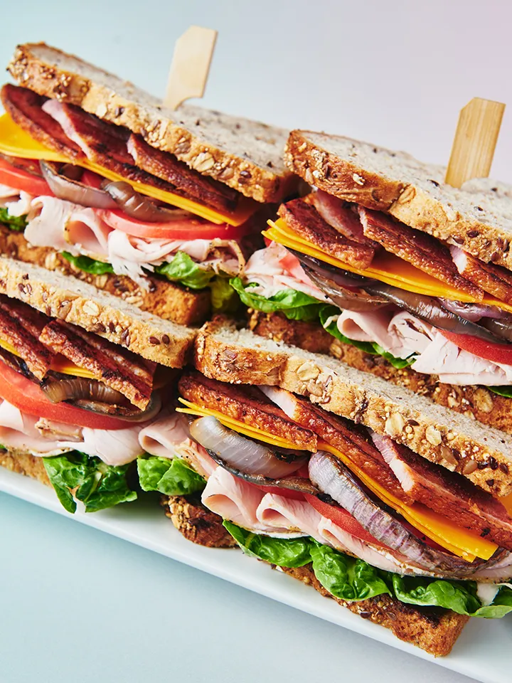 Four stacked turkey bacon cheddar sandwiches on multigrain bread with lettuce, tomato, and grilled onions.