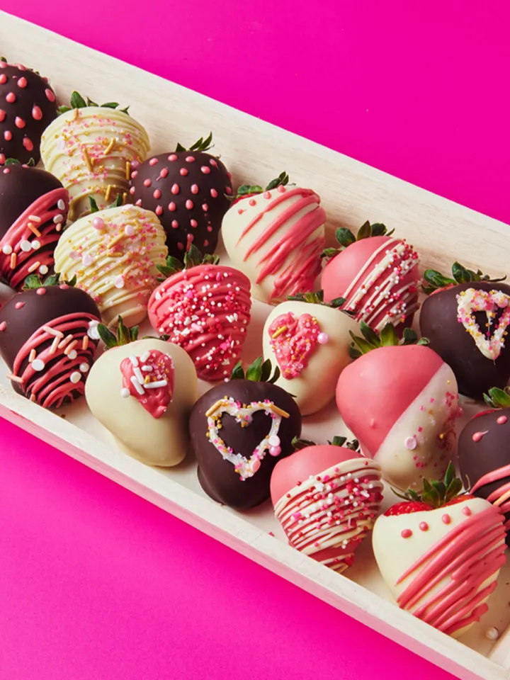 Assorted strawberries dipped in white, dark, and pink chocolate with decorative sprinkles on a wooden tray.