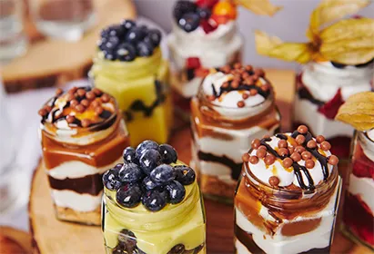Jars with layered desserts topped with blueberries, cream, chocolate drizzle, and crunchy toppings on a wooden board.