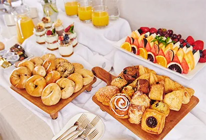 Buffet table with bagels, assorted pastries, fresh fruit slices, parfaits, and glasses of orange juice.