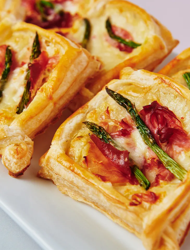 Baked puff pastry squares topped with asparagus spears, prosciutto, and melted cheese.