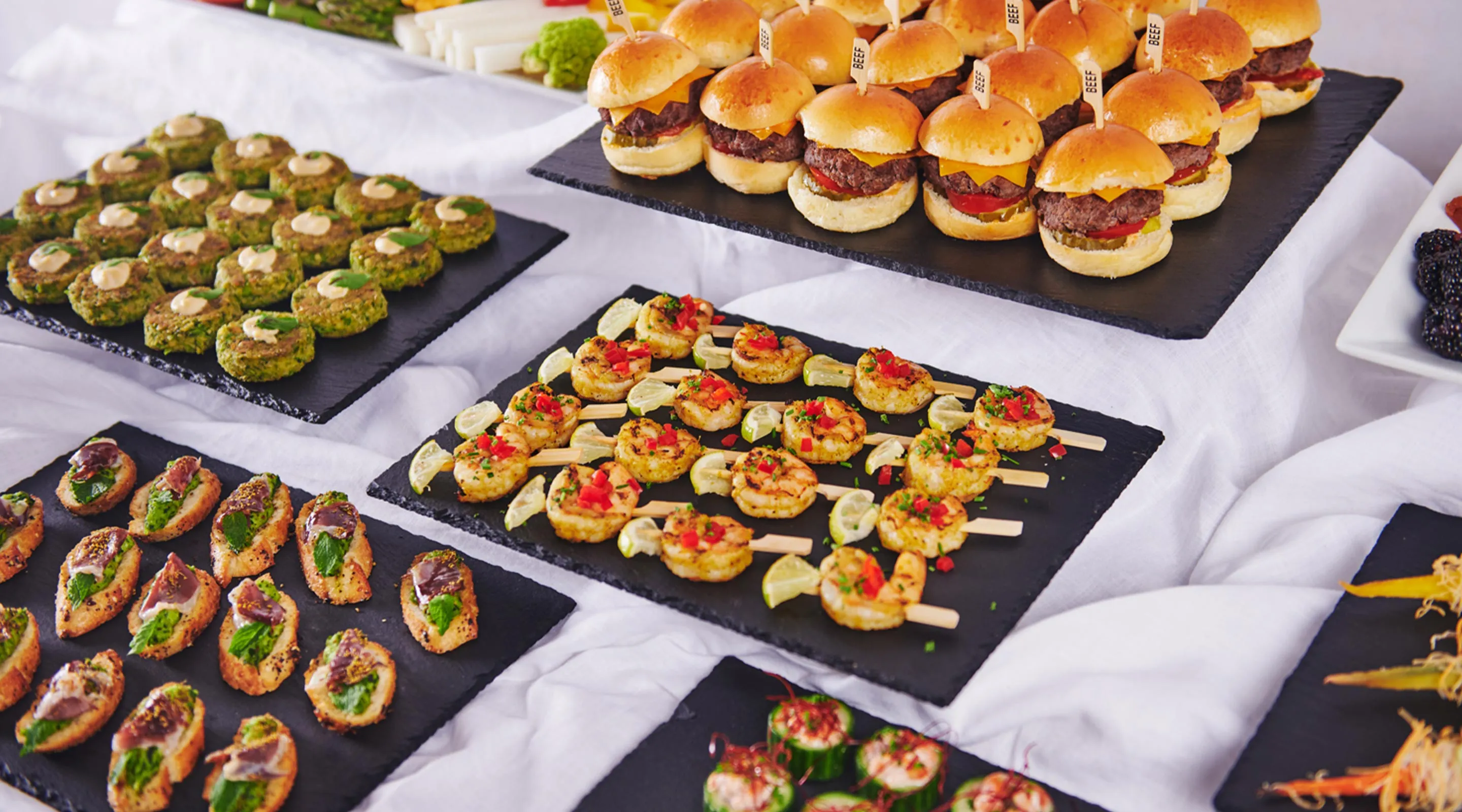 Plates of assorted appetizers including mini beef burgers, shrimp skewers, and crostini on a white tablecloth.
