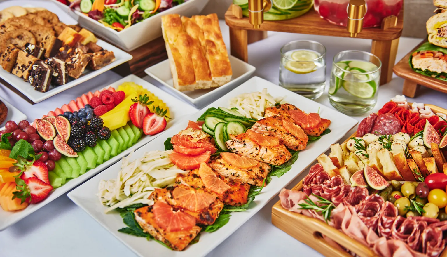 Platter with grilled salmon, fresh fruit, charcuterie, bread, salad, and lemon water on a white tablecloth.