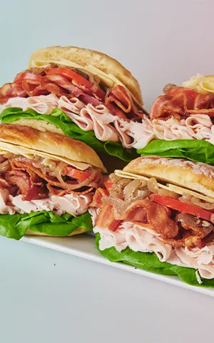 Three sandwiches with turkey, Irish cheddar, bacon, lettuce, tomato, and onions on a white plate.