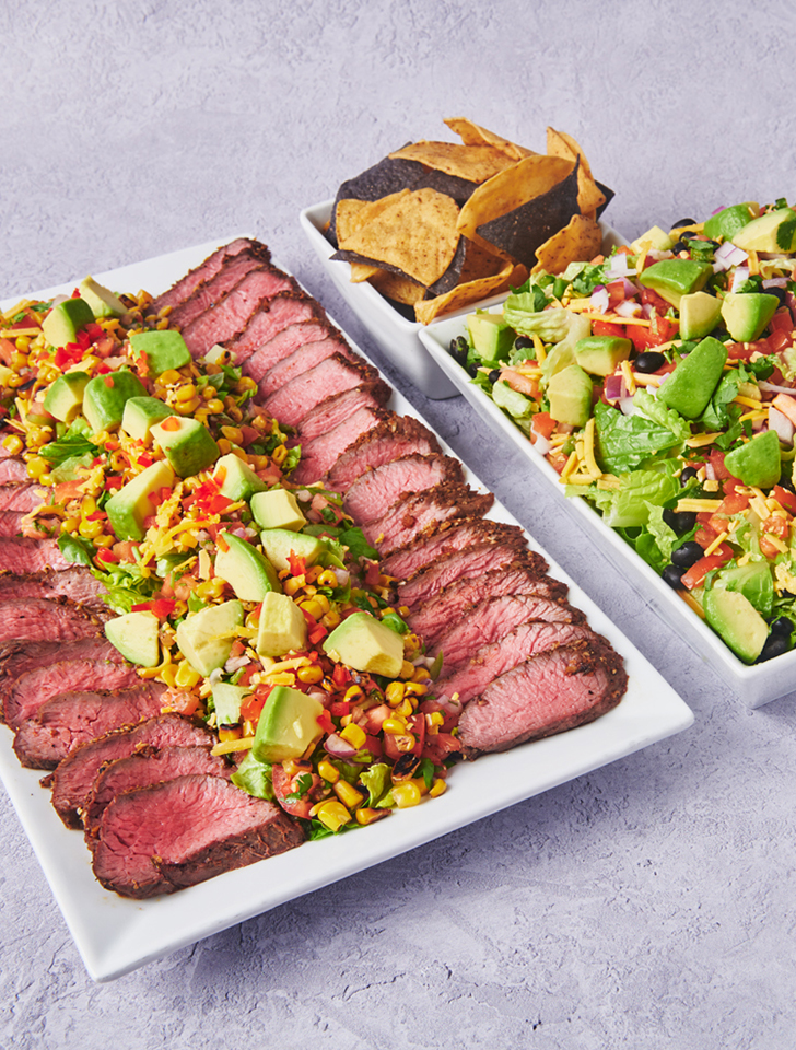 Catering platter with sliced steak, avocado, corn salsa, tortilla chips, and a fresh Southwestern-style salad.