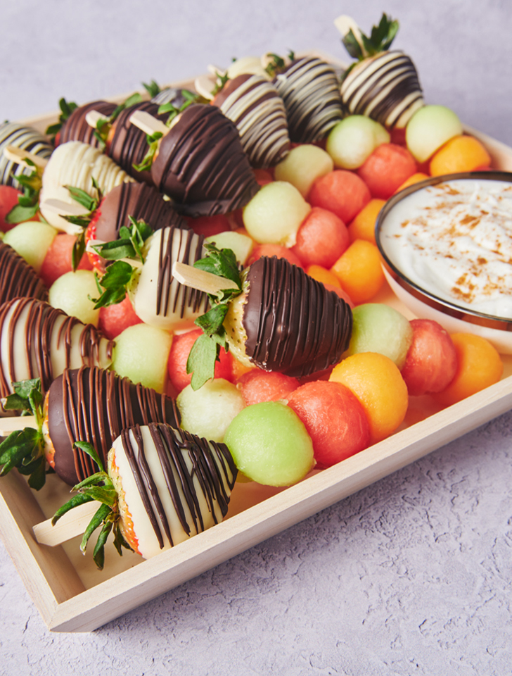 Chocolate-dipped strawberries with melon balls and a creamy fruit dip served on a wooden catering tray.