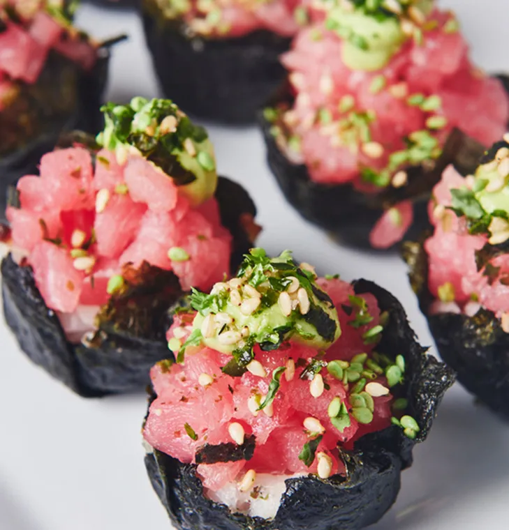 Close-up of sushi topped with diced raw tuna, green herbs, sesame seeds, and seaweed wraps.
