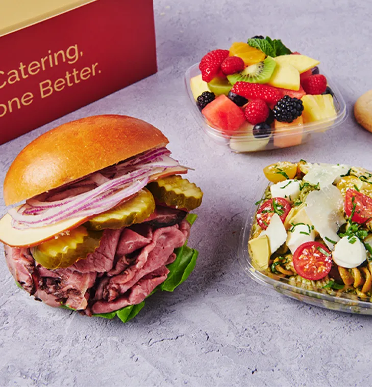 A roast beef sandwich with pickles and onions, a fruit salad, and a pasta salad on a gray surface.