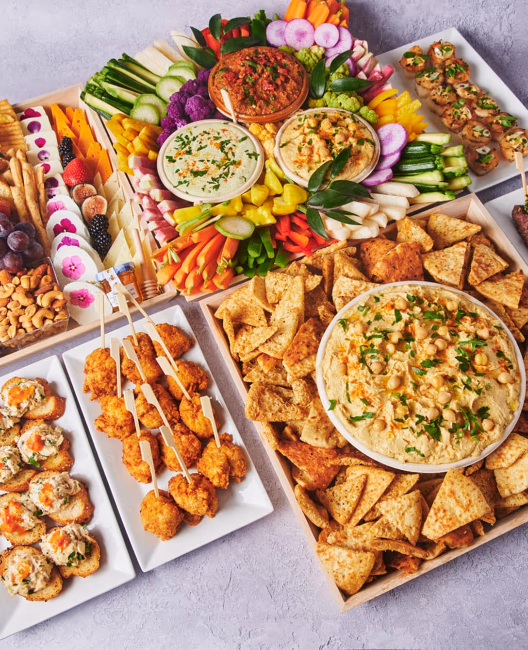 Assorted catering package with crudités, dips, pita chips, fruit, cheese, skewers, and bite-sized appetizers arranged on serving trays.