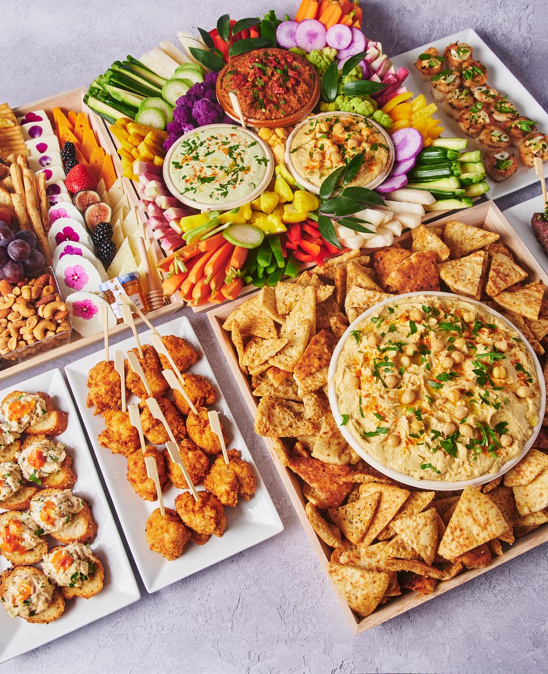 Assorted catering package with crudités, dips, pita chips, fruit, cheese, skewers, and bite-sized appetizers arranged on serving trays.