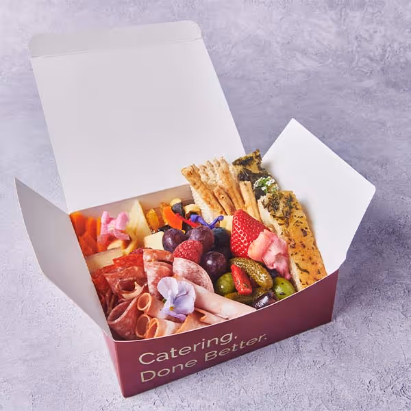Branded catering box filled with charcuterie, fruit, cheese, crackers, and vegetables.