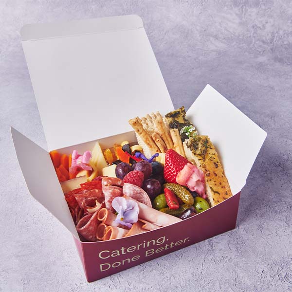Branded catering box filled with charcuterie, fruit, cheese, crackers, and vegetables.