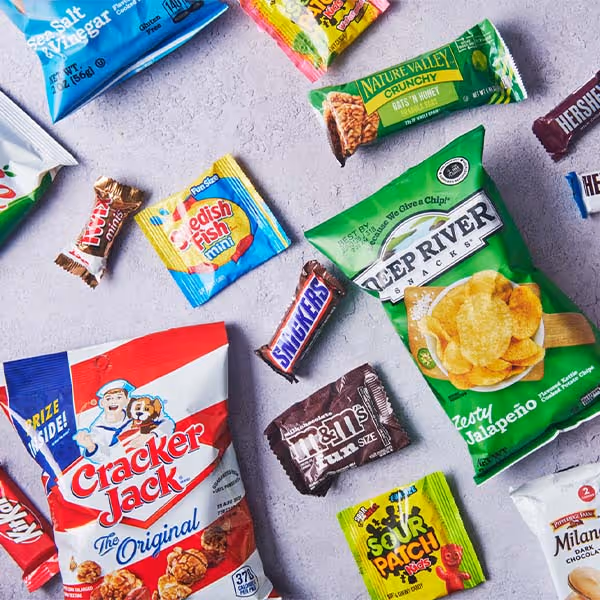 Assorted packaged snacks, chips, candy, and Cracker Jack arranged on a light gray surface.