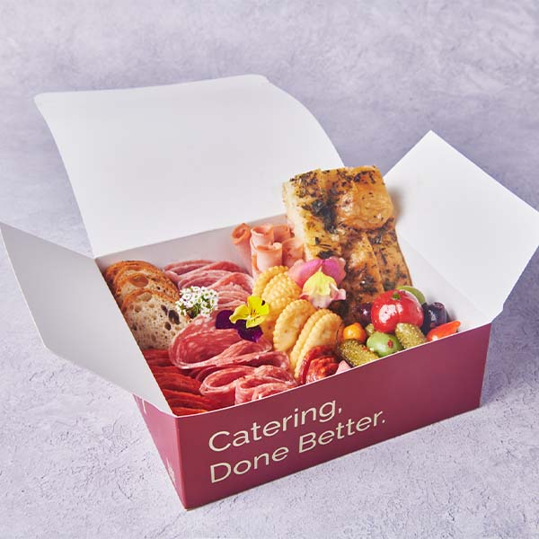 Branded catering box filled with meats, cheese, bread, fruit, and fresh vegetables.