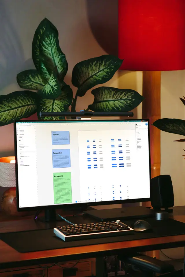Desktop monitor showing The Project Launchpad design system in Figma, displayed in a warm studio workspace with plants, keyboard, and lighting.