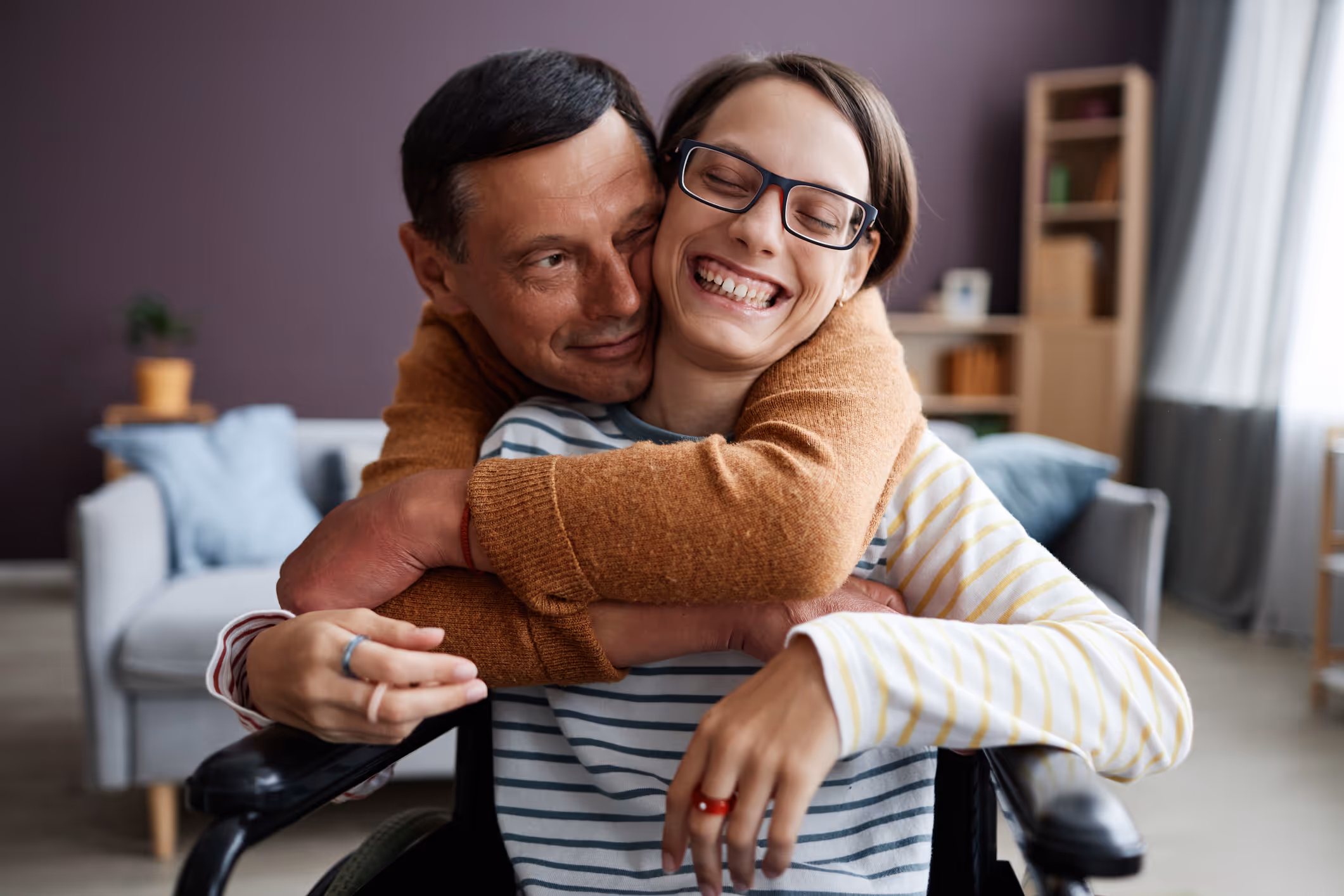 Smiling woman in wheelchair embraced by her partner at home, sharing a joyful and supportive moment together