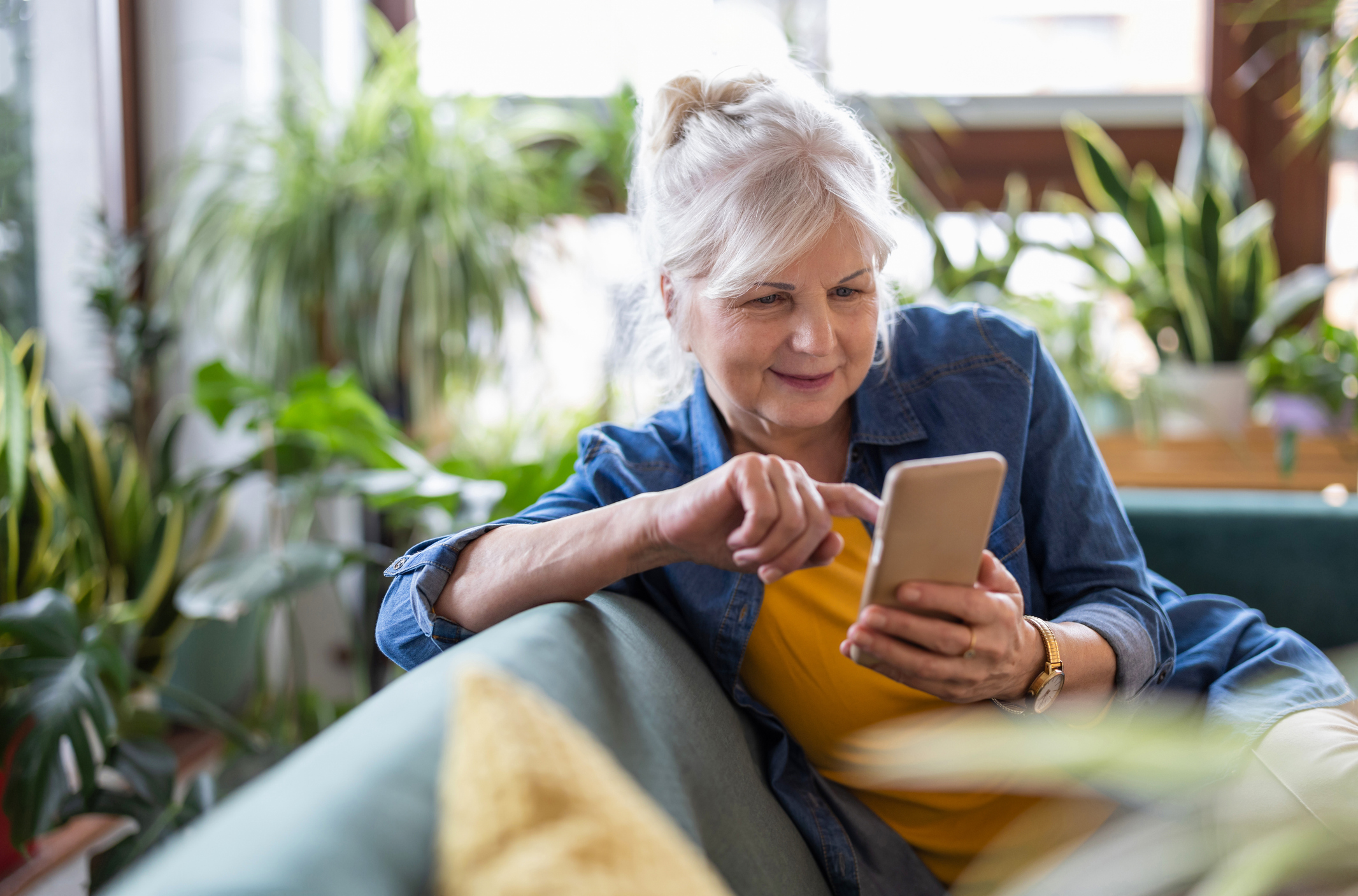 Older woman relaxing at home on couch, smiling while using smartphone to browse or connect online