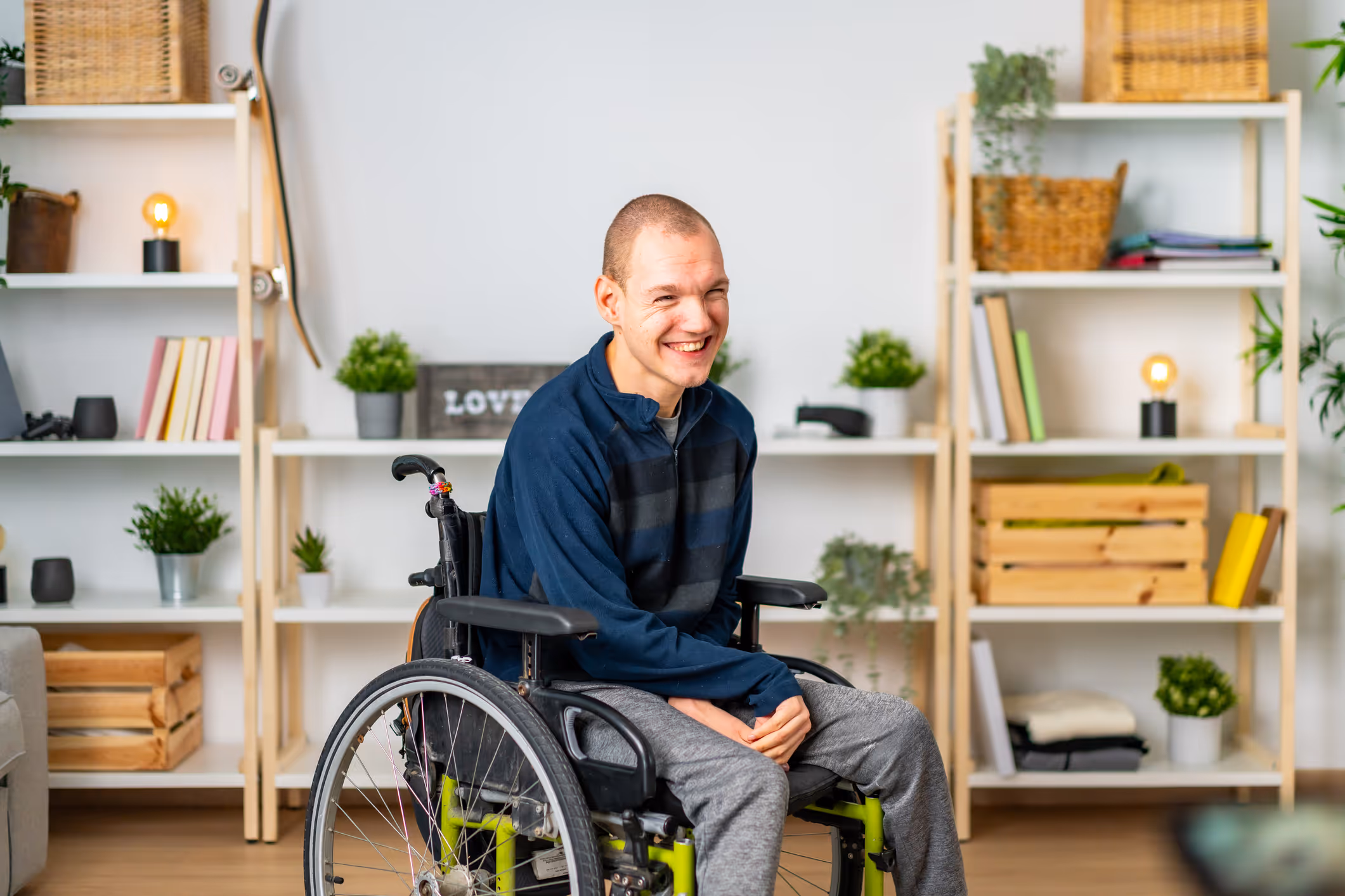 Man with disability smiling while seated in wheelchair at home, surrounded by plants and personal items