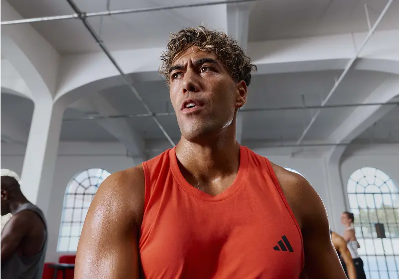 One person in a red Adidas sleeveless shirt stands in a gym with white walls and arched windows; three others are in the background, including one lifting weights in a Les Mills Bodypump workout.