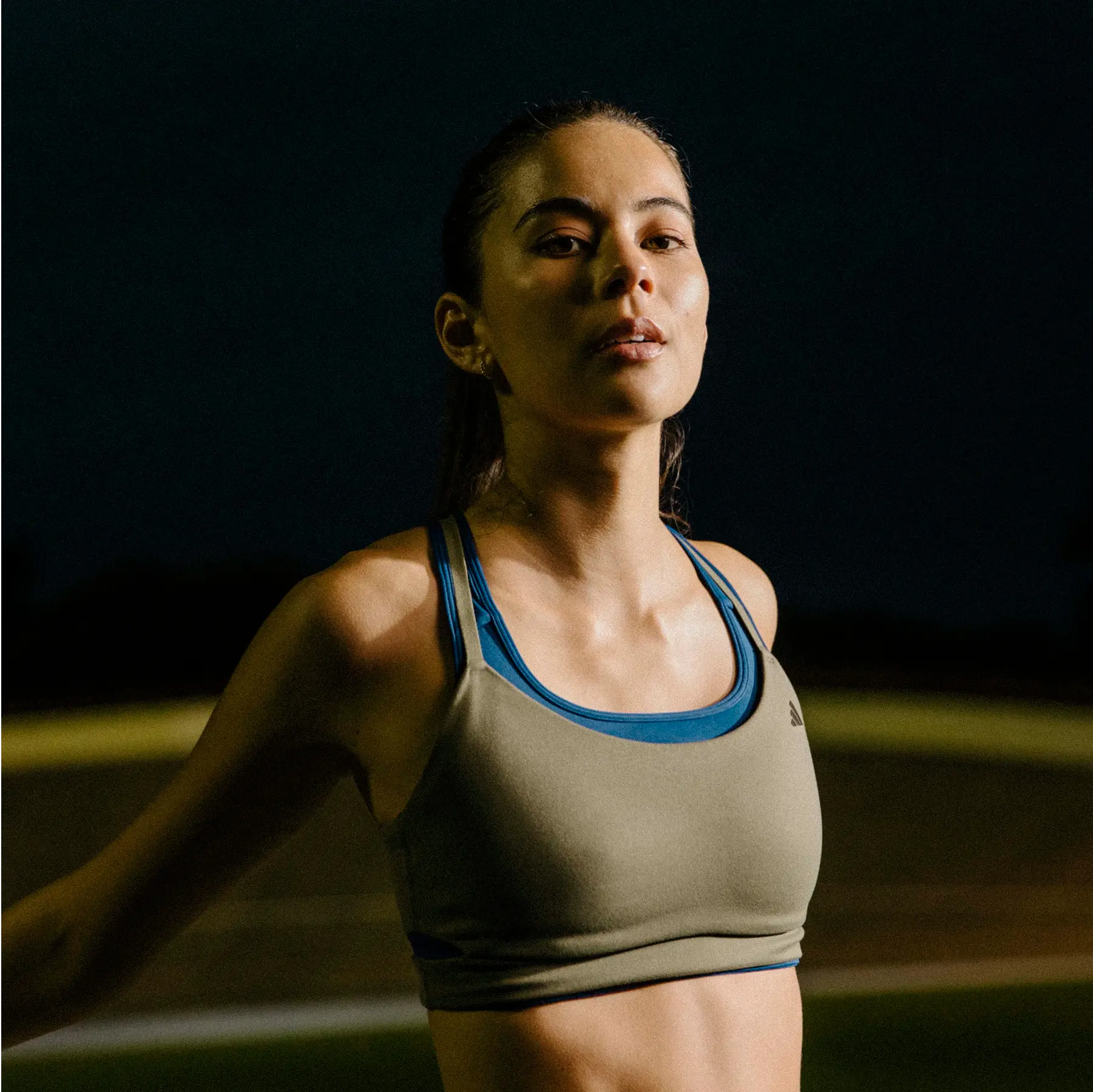 One person with a blurred face stands in a gymt, wearing a grey sports bra with blue straps and blue athletic shorts after doing a Les Mills Grit workout. The background is dark and indistinct.