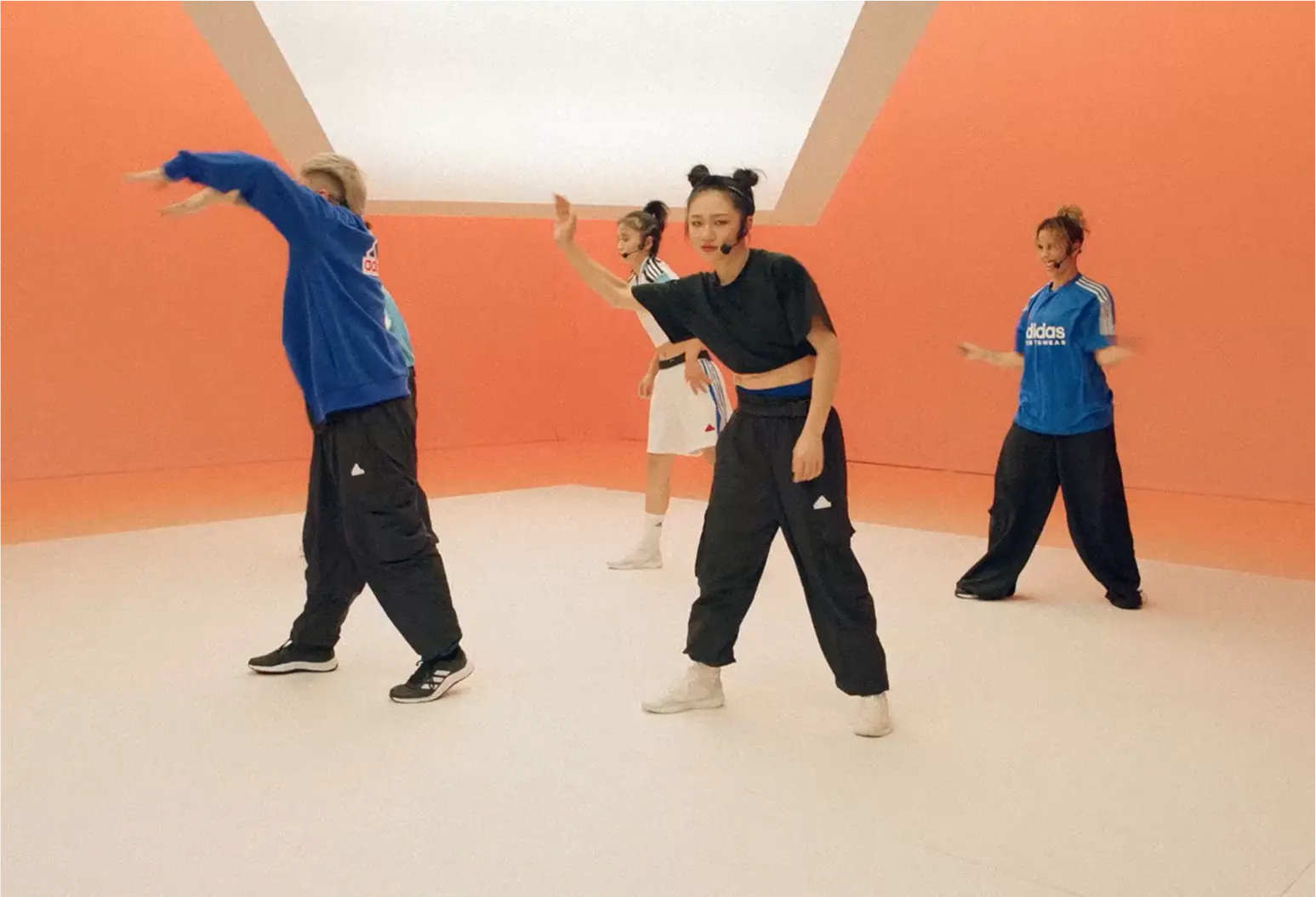 Four people in athletic wear dancing in a studio with an orange wall and white floor, each wearing a headset microphone.