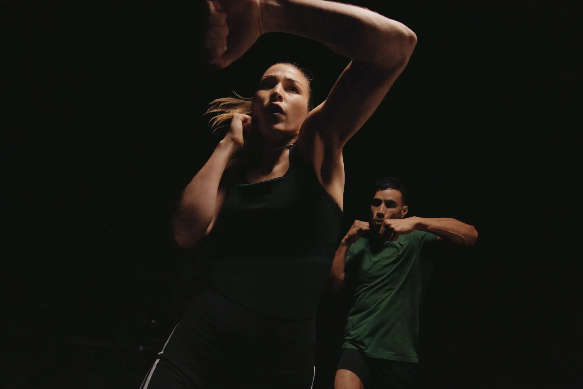 Two people practice boxing in a dark room - one in a black tank top in the foreground, the other in a green shirt in the background - both with arms raised.