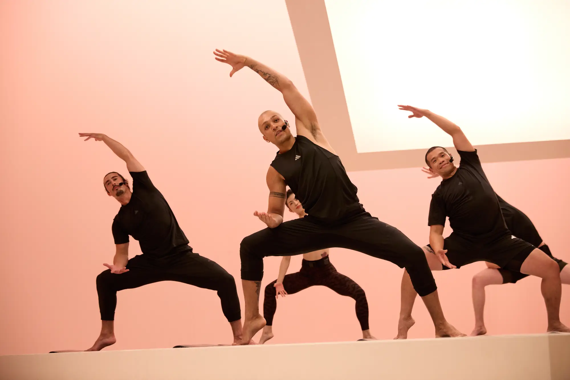 Five people in black athletic wear perform a BodyBalance pose on a light pink background, standing with legs apart, one arm extended overhead and the other bent at the elbow.