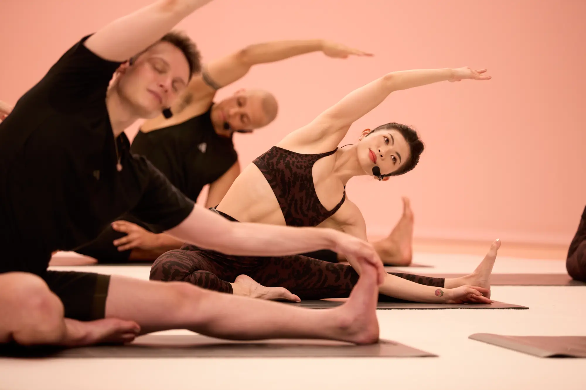 Three people in black athletic wear perform a BodyBalance pose on a light pink background, sitting on the ground and stretching sideways.