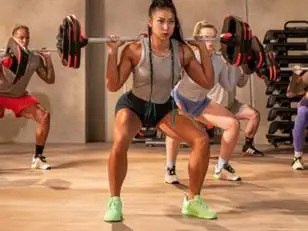 Four people in a gym perform barbell squats with black and red weight plates. The front person wears a gray top, black shorts, and neon green shoes; others wear athletic outfits in neutral tones.