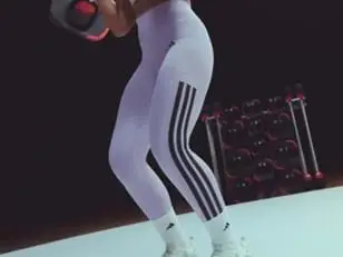 One person wearing white leggings with black stripes and white shoes holds a black and red weight plate on a white platform. A rack of similar weights is visible in the background.