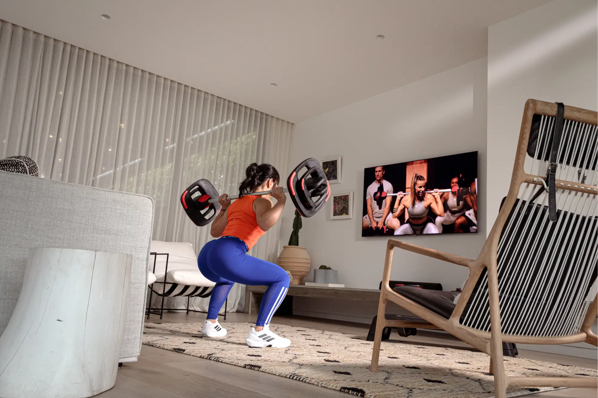 One person in orange top and blue leggings doing a squat with a barbell in a bright living room. A TV shows a workout video with three instructors in white and red outfits.