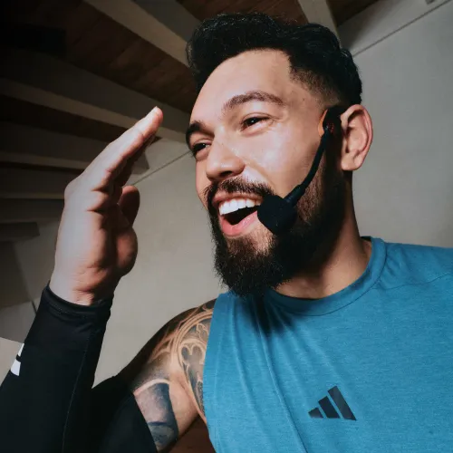 Smiling man with a beard wearing a headset microphone and a blue Adidas sleeveless shirt, raising his hand indoors.