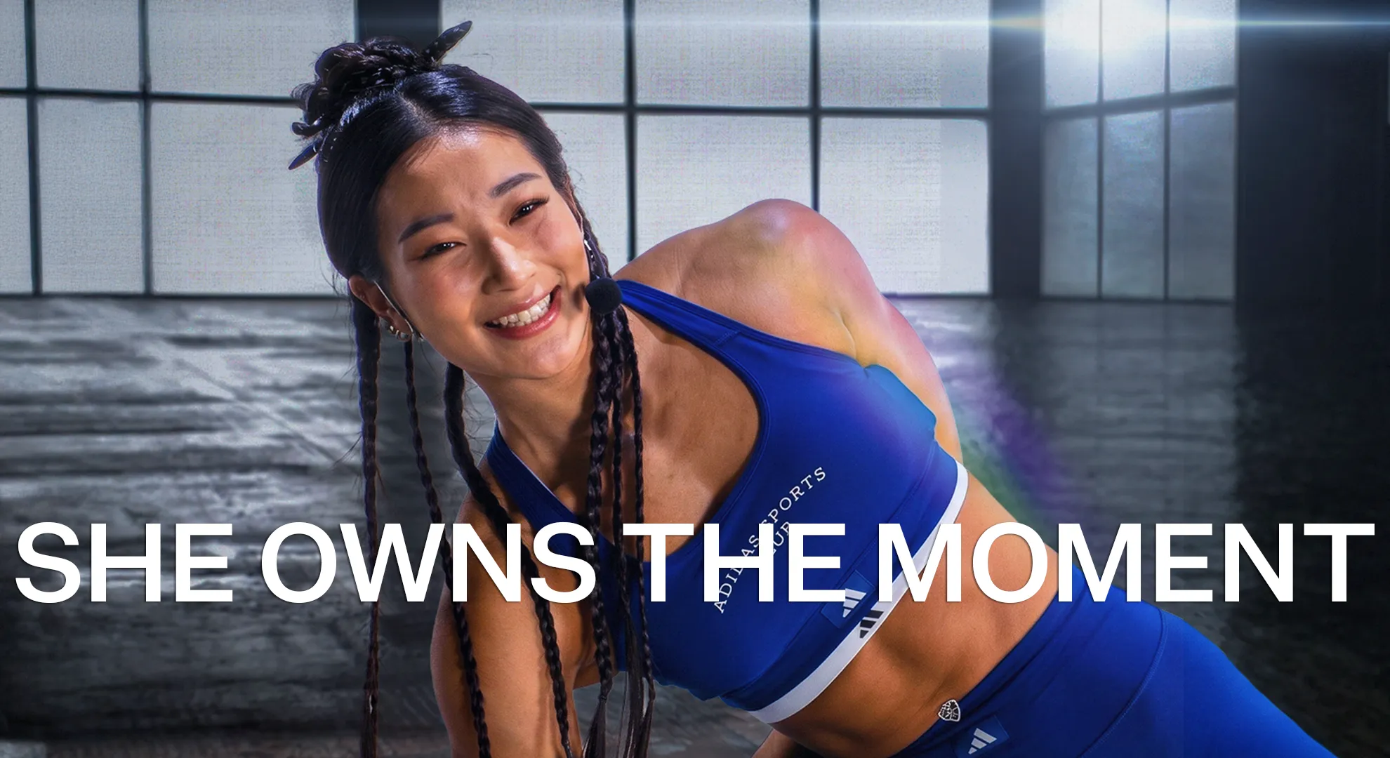 Smiling woman in blue athletic wear doing a side plank exercise in a gym with large windows.