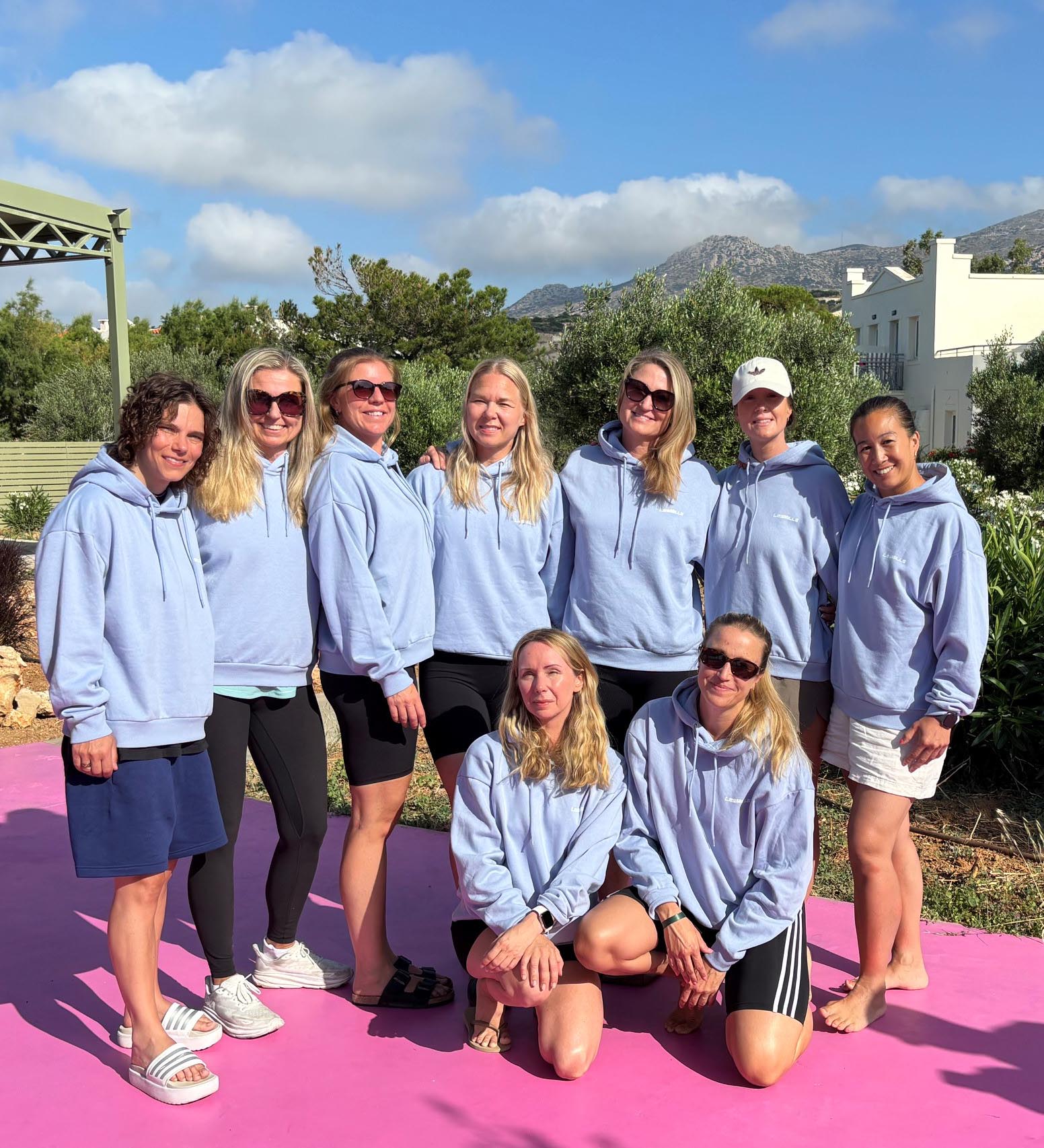 Group of women wearing light blue hoodies posing outdoors on a pink floor with trees and mountains behind.