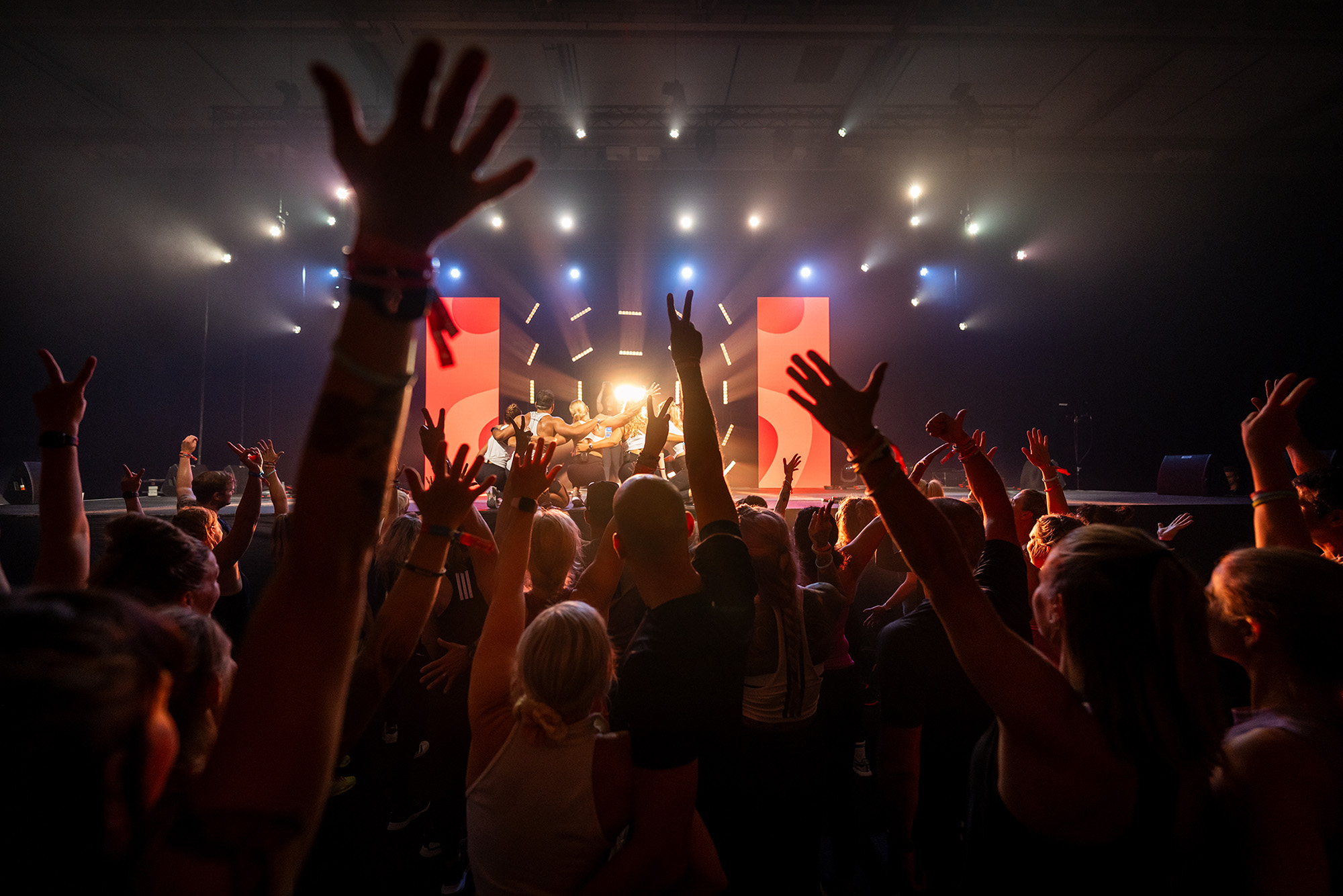 Crowd with raised hands facing a brightly lit stage with performers in front of red panels.