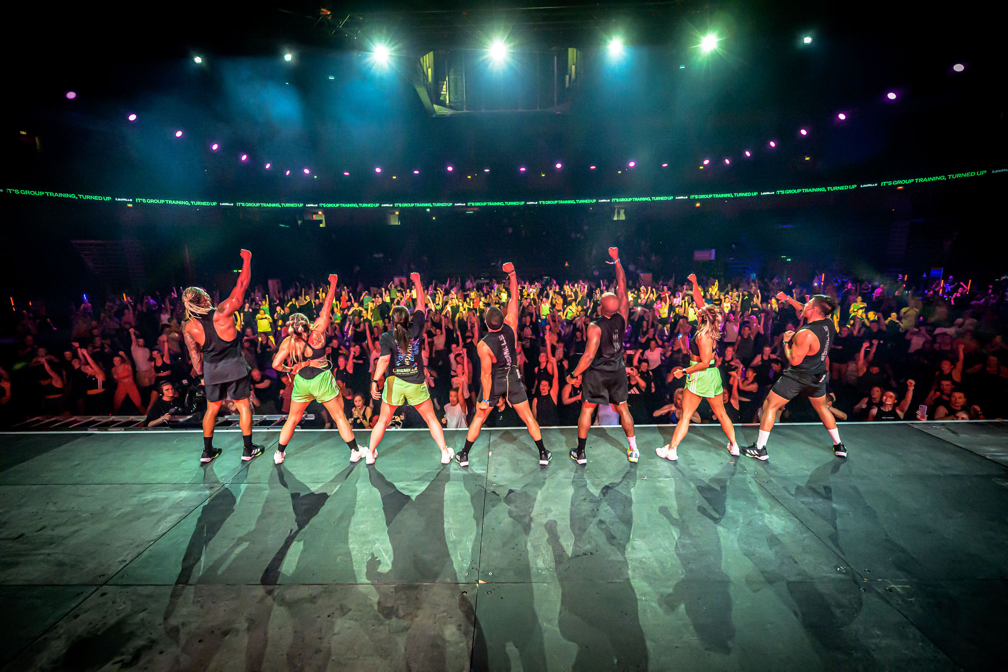 Seven fitness instructors on stage with raised arms facing a large cheering crowd under spotlights.