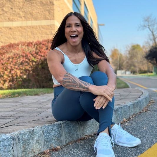Image of Lula Slaughter sitting on a footpath curb with arms crossed and smiling at the camera